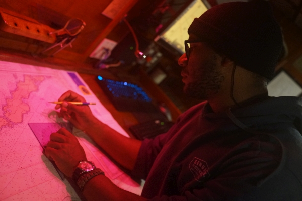 Picture shows a student belowdecks on a ship sitting at a desk covered in a nautical chart, pencil and protractor in hand. It's nighttime, and the lighting is dim and red to preserve night vision
