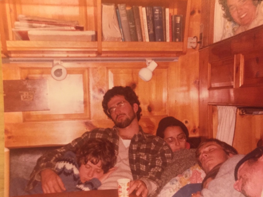 Image shows three college students slumped side by side, napping in a cozy, wood-paneled nook belowdecks, with two bunks just visible in a wall to the right